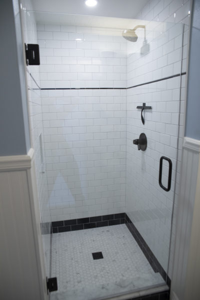 A modern walk-in shower featuring a frameless glass door and white subway-tiled walls. The shower floor is finished with white and black penny tile, while dark oil-rubbed bronze fixtures provide a sharp contrast.
