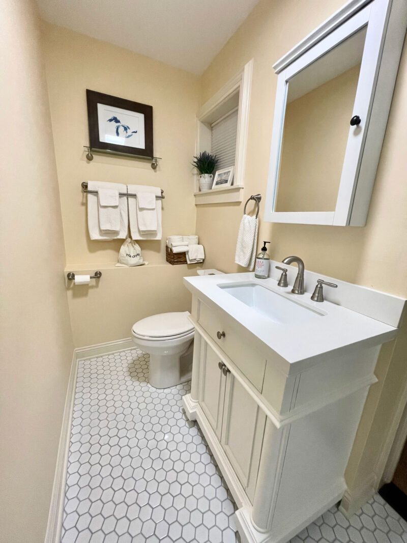 A narrow bathroom with cream walls and white honeycomb floor tiling. The space features a white vanity with a rectangular sink and brushed nickel fixtures, a white toilet, and a medicine cabinet with a mirrored door. Neatly folded white towels hang on a rack below a framed piece of wall art.