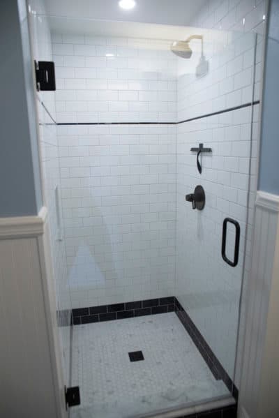 A modern walk-in shower featuring a frameless glass door and white subway-tiled walls. The shower floor is finished with white and black penny tile, while dark oil-rubbed bronze fixtures provide a sharp contrast.