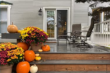 A wooden deck decorated for autumn with various orange pumpkins and large pots of yellow and purple chrysanthemums. The steps lead up to a gray house with a sliding glass door and two patio chairs.