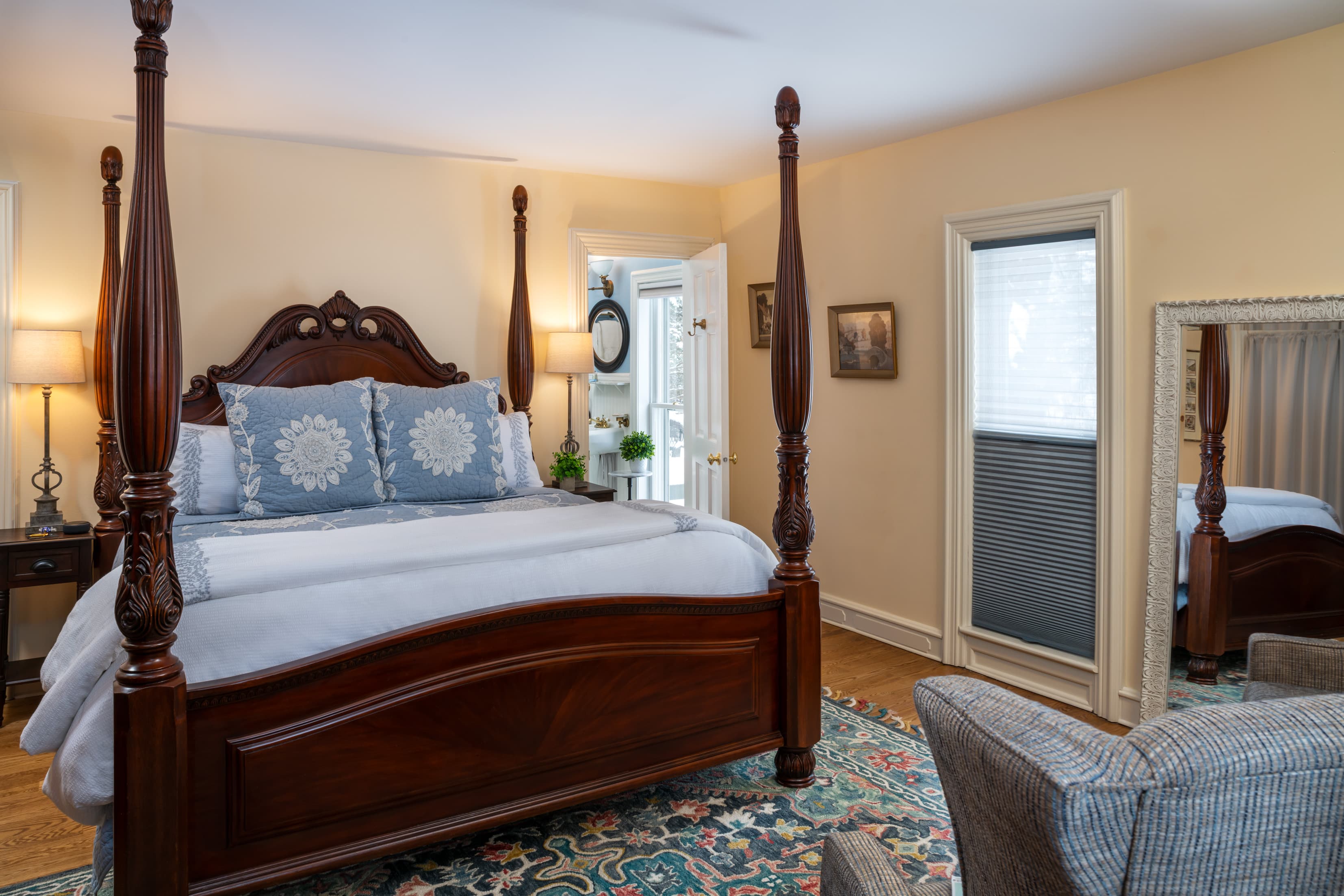 A traditional bedroom featuring a dark wood king four-poster bed with a carved headboard, light blue bedding, and decorative blue pillows. The room includes a patterned area rug, a full-length floor mirror, and warm beige walls.