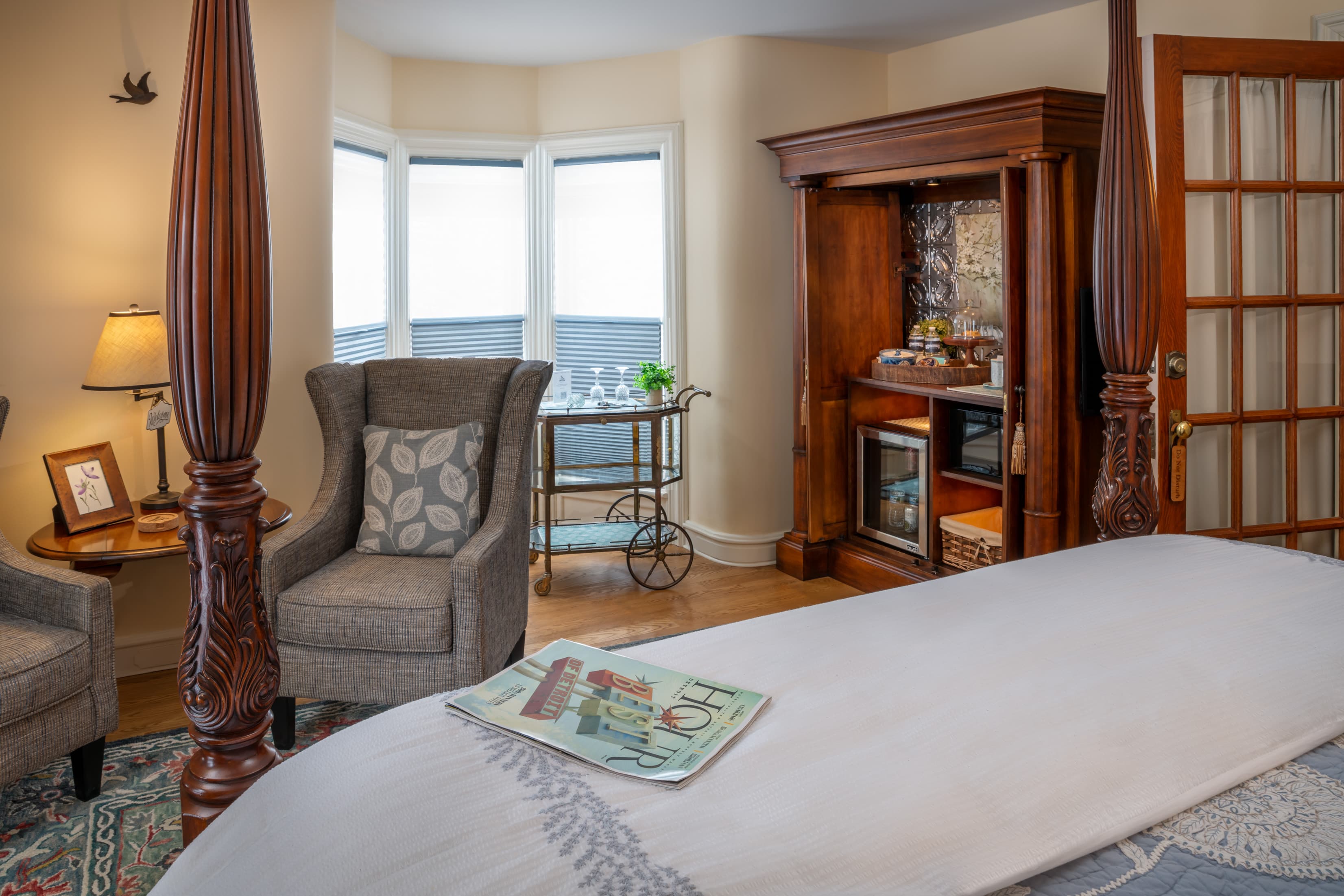 A cozy bedroom nook featuring a gray wingback chair with a leaf-patterned pillow next to a bay window. The space includes a wooden armoire repurposed as a coffee station with a mini-fridge, a gold bar cart, and a glimpse of a four-poster bed in the foreground.