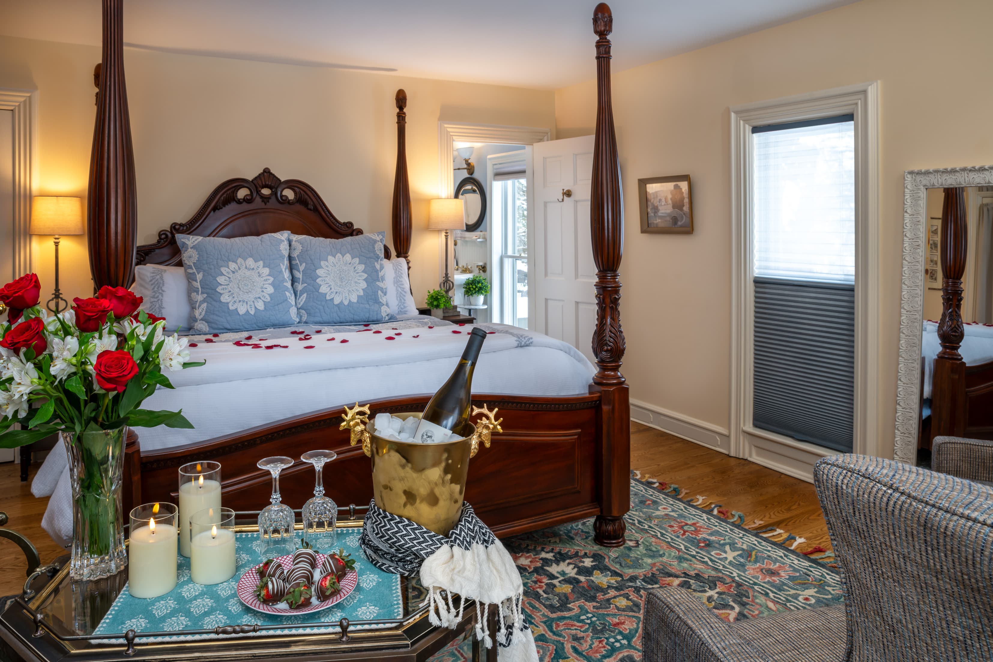 A romantic bedroom setting featuring a dark wood four-poster bed sprinkled with red rose petals. In the foreground, a tray holds a bottle of champagne in an ice bucket, two glasses, lit candles, chocolate-covered strawberries, and a vase of red roses.