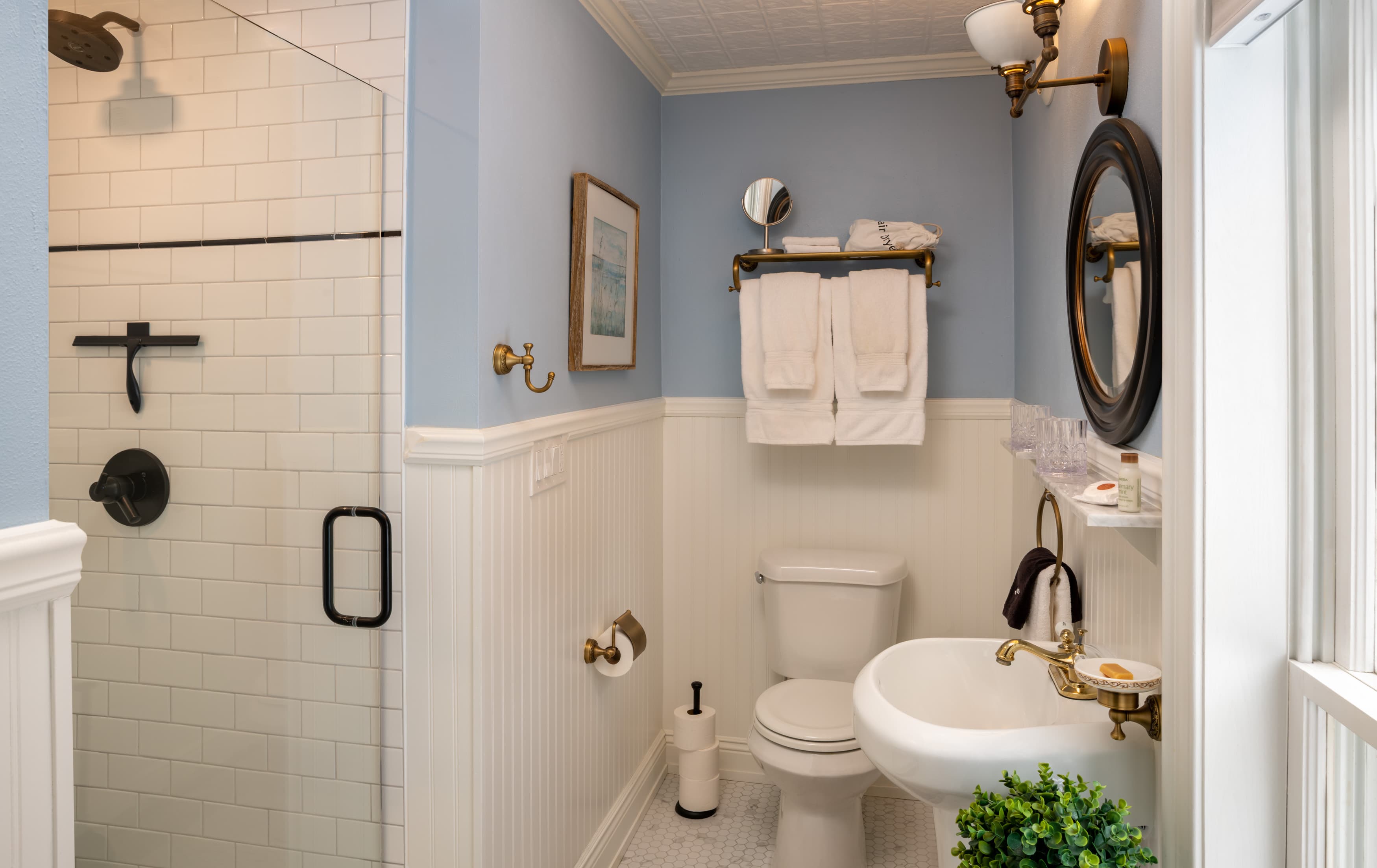 A bright, modern bathroom featuring light blue walls and white wainscoting. The space includes a glass-enclosed walk-in shower with white subway tiles, a white pedestal sink with brass fixtures, a round mirror, and neatly folded white towels on a brass rack.