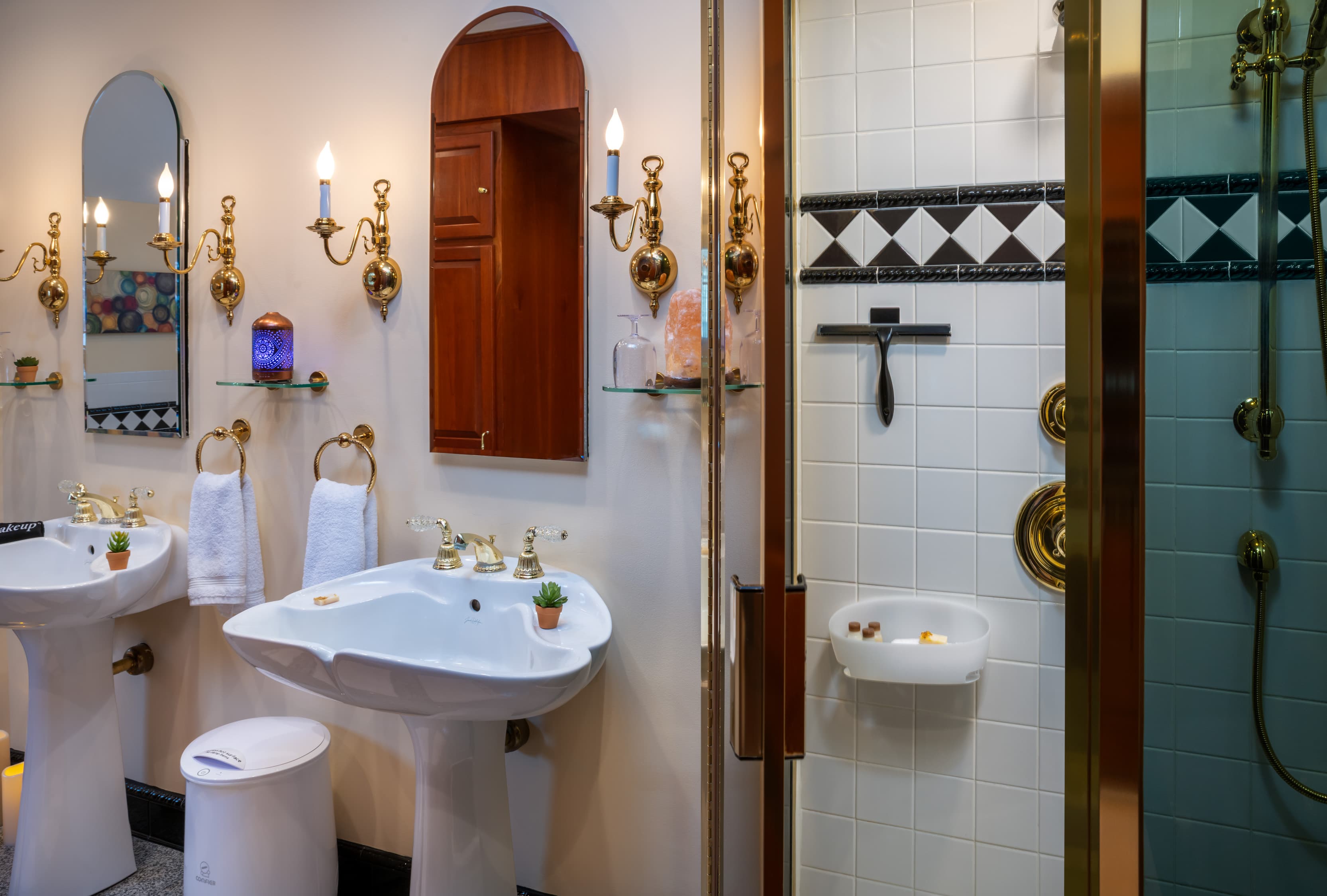A brightly lit, vintage-style bathroom featuring two white pedestal sinks with gold faucets and hardware. Above each sink is an arched mirror flanked by gold candle-style wall sconces. To the right, a glass-enclosed shower is visible, featuring white wall tiles with a black-and-white diamond border and additional gold fixtures.