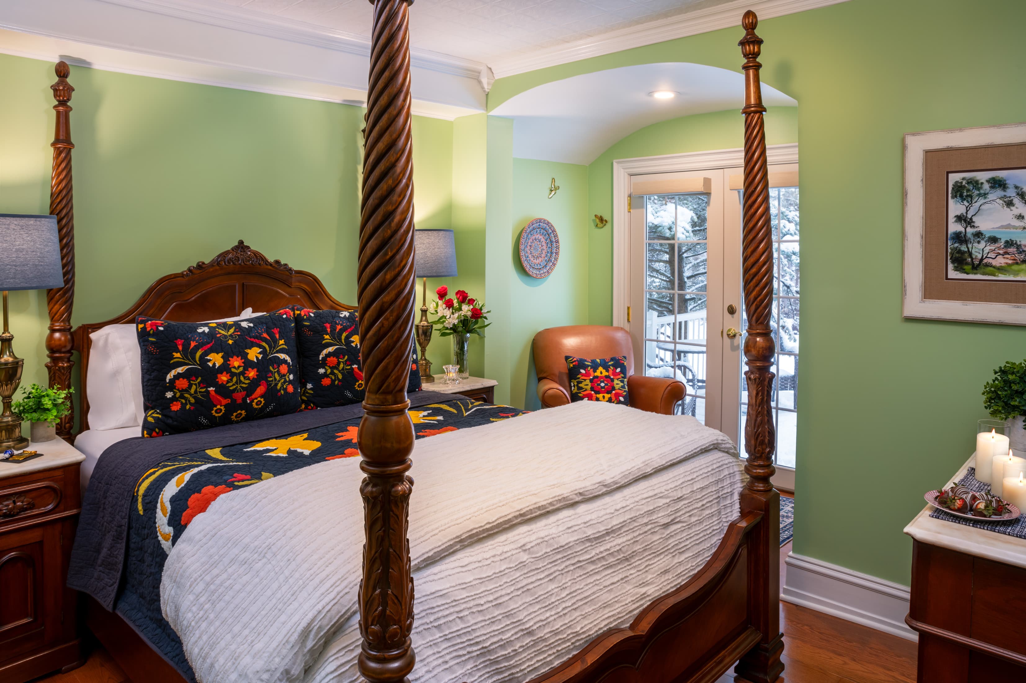 A vibrant bedroom with sage green walls and a wooden four-poster bed. The bed features a white quilt and dark blue pillows with colorful floral embroidery. In the background, a tan leather armchair sits near a set of glass doors leading to a snowy outdoor view.