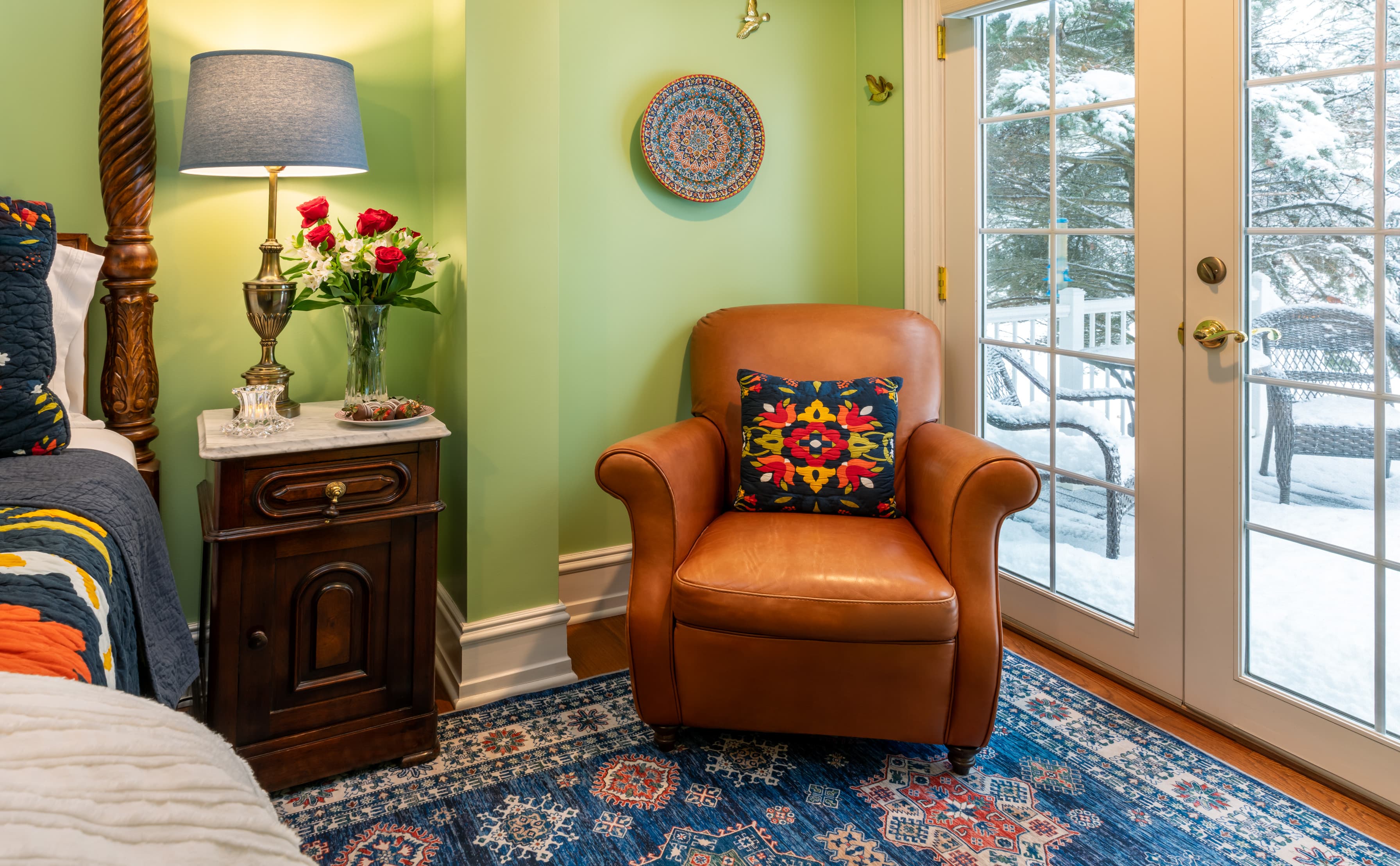 A cozy reading corner in a bedroom with sage green walls, featuring a tan leather armchair with a vibrant, embroidered floral pillow. The space is set on a patterned blue area rug next to glass French doors that look out onto a snowy deck.