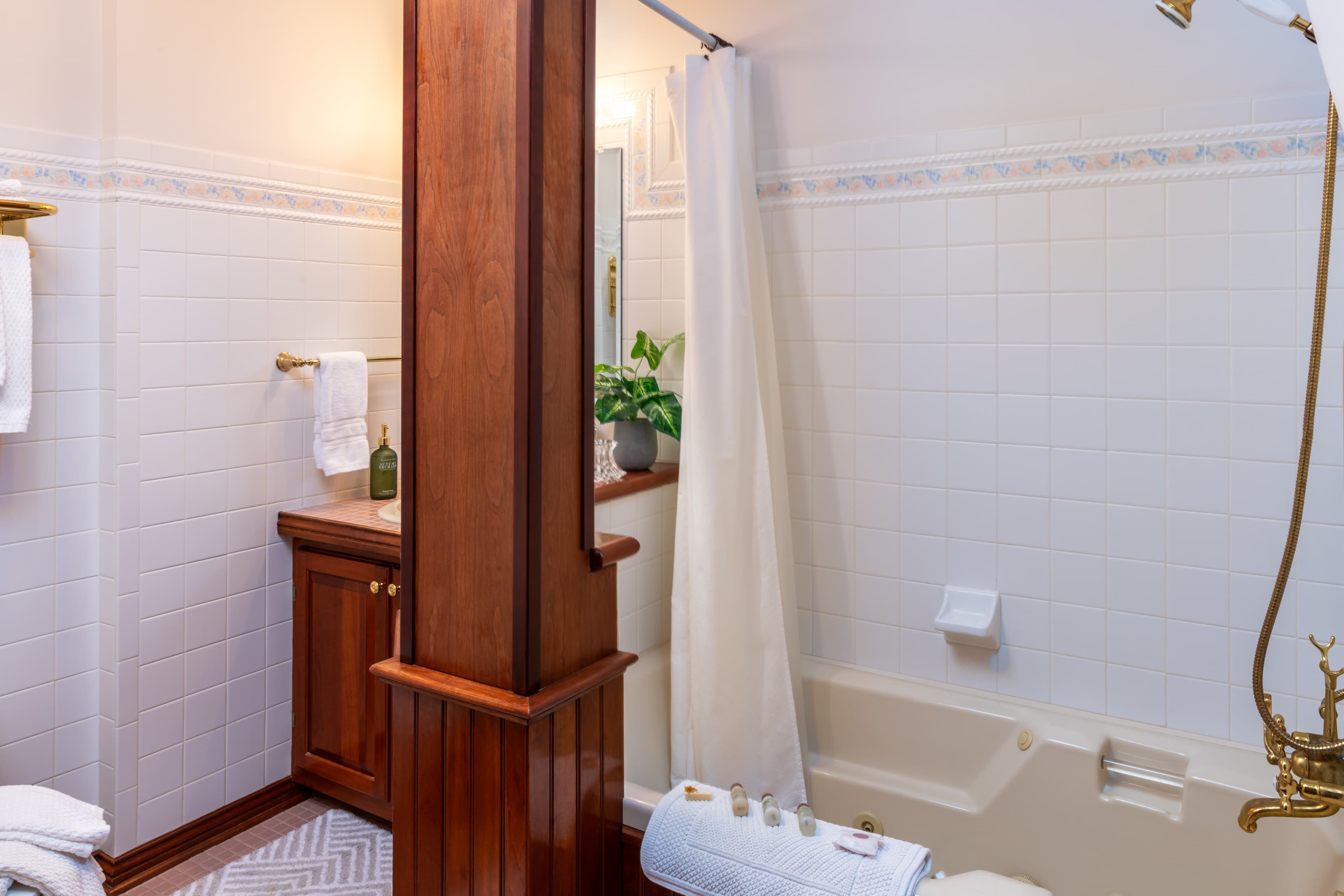 A classic bathroom featuring white wall tiles and a beige bathtub with gold fixtures. A prominent dark wood pillar stands in the foreground, partially obscuring a matching wood vanity with a white sink. A white shower curtain is pulled to the side, and a small green plant sits on the ledge behind the pillar.