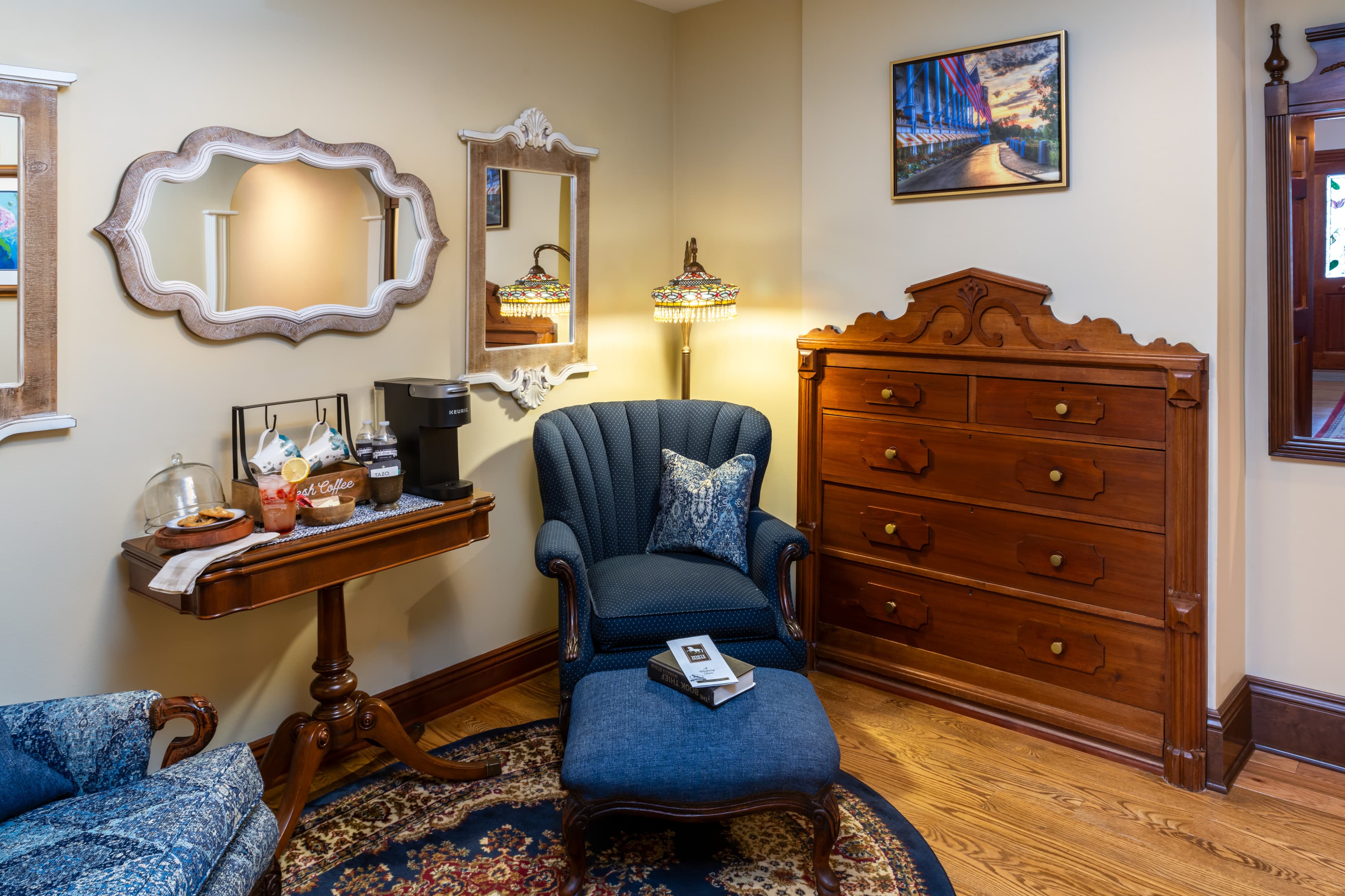 A cozy sitting area in a bedroom featuring a navy blue scalloped armchair and matching ottoman with a small book on top. The room includes a tall dark wood dresser, a small pedestal table with a coffee maker and snacks, and two uniquely shaped white-framed mirrors on the beige wall.