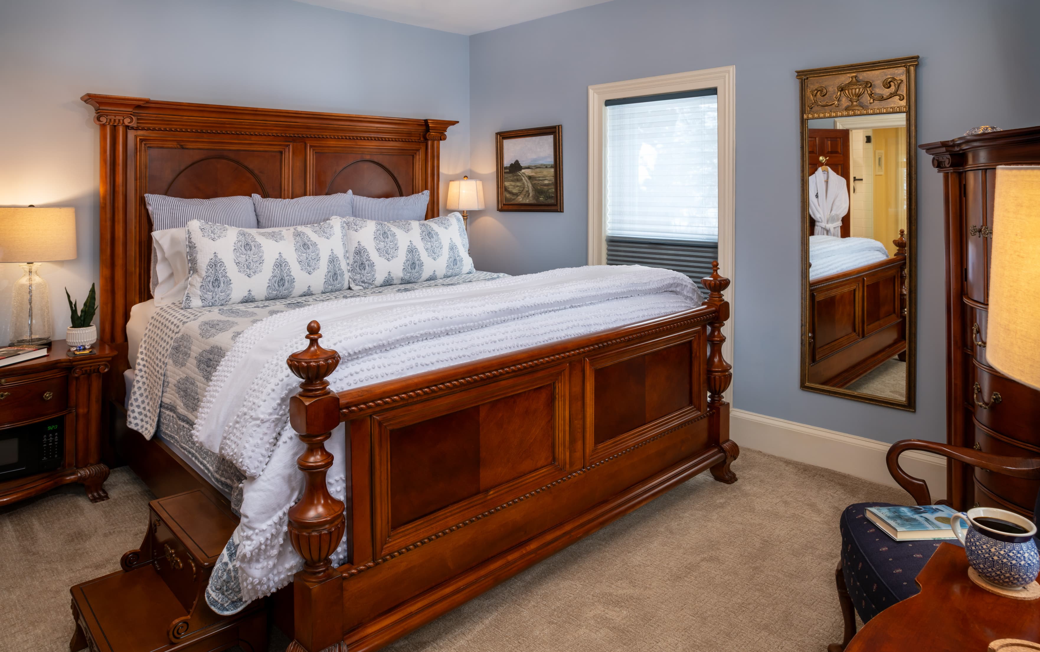 A spacious bedroom featuring a high, dark wood king bed with a large headboard and footboard, dressed in light gray bedding and patterned pillows. The room has light blue walls, a full-length mirror reflecting a white bathrobe, and a small set of wooden steps leading up to the side of the bed.
