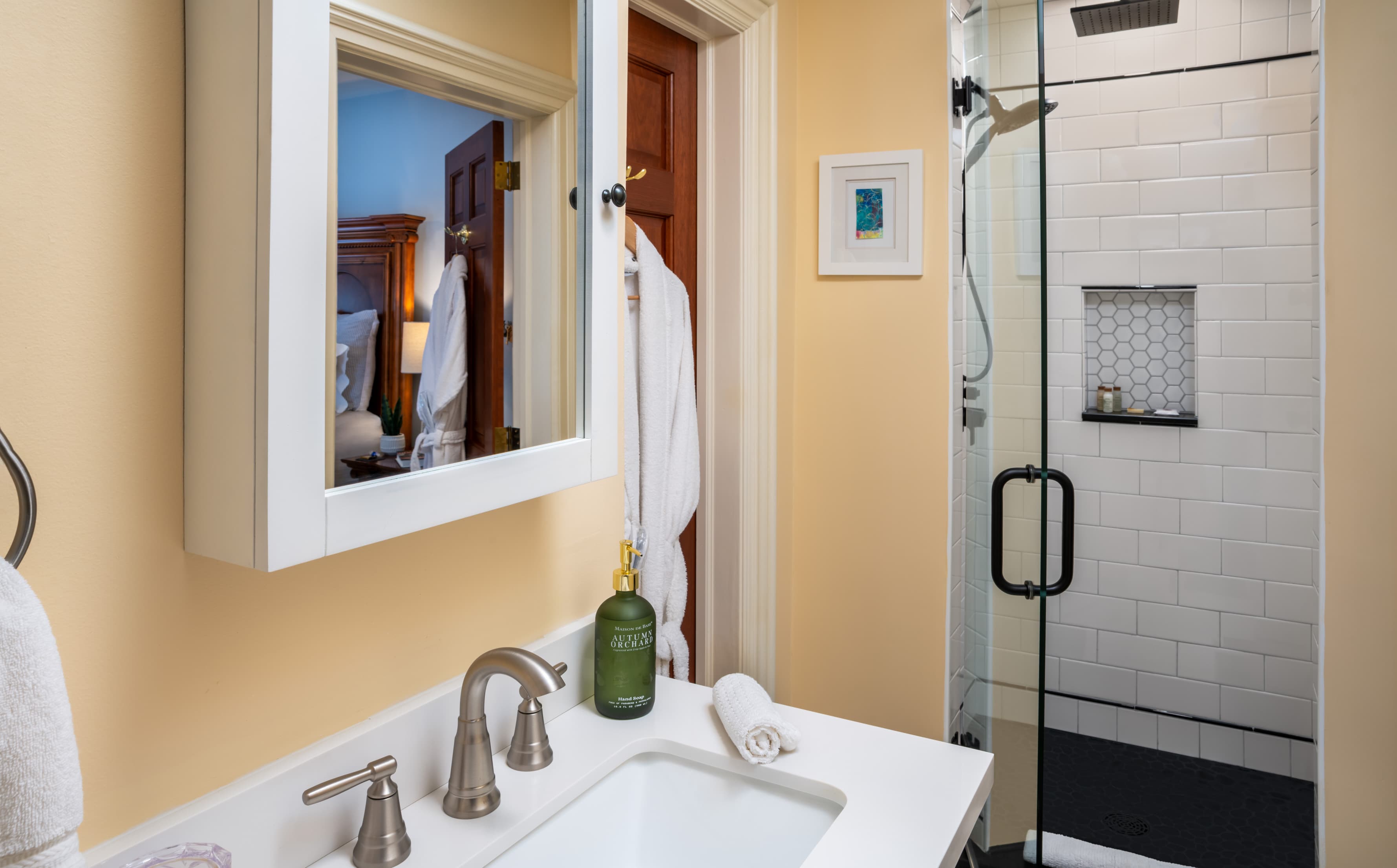 A bright bathroom featuring a white vanity with a rectangular sink and brushed nickel fixtures. In the background, a glass-enclosed walk-in shower with white subway tiles and a recessed shelf is visible next to a white bathrobe hanging on a wooden door.