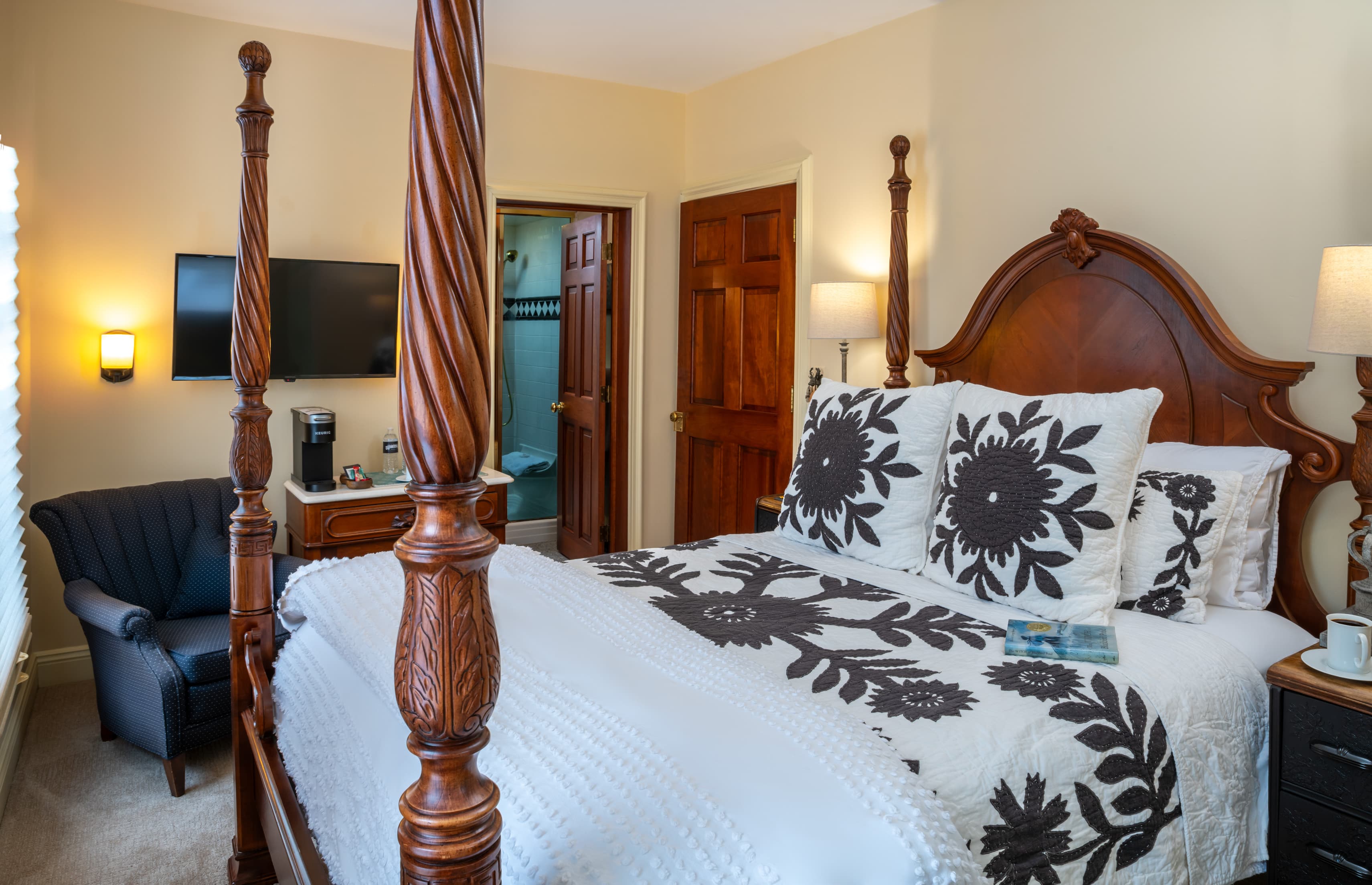 A classic bedroom featuring a wooden four-poster bed with white bedding and gray floral-patterned pillows. The room includes a navy blue armchair in the corner, a wall-mounted television, and a doorway leading to a brightly lit bathroom.
