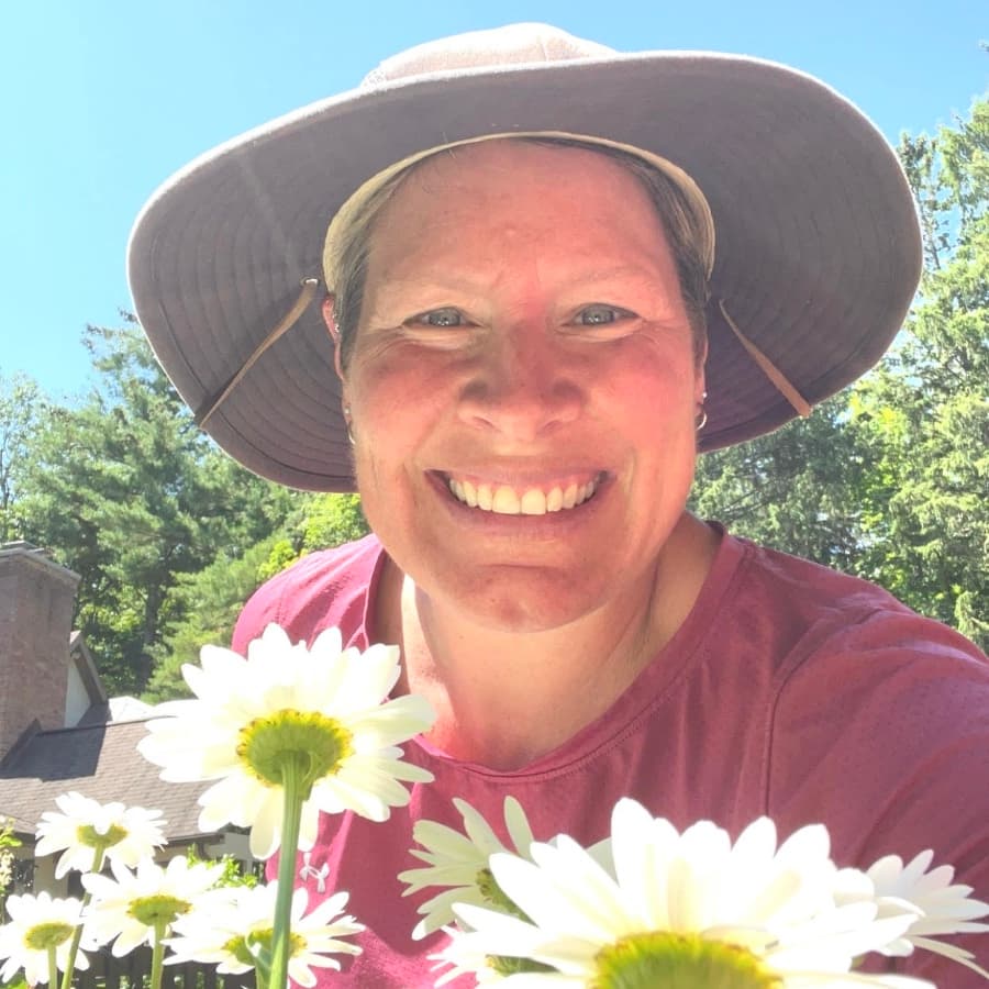 A smiling person in a wide-brimmed hat poses among white daisies under a clear blue sky.