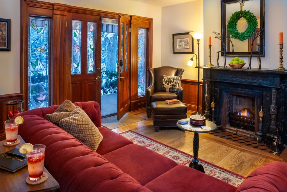 A warm, cozy living room featuring a red sofa, a fireplace, and large front doors leading to a winter scene outside.