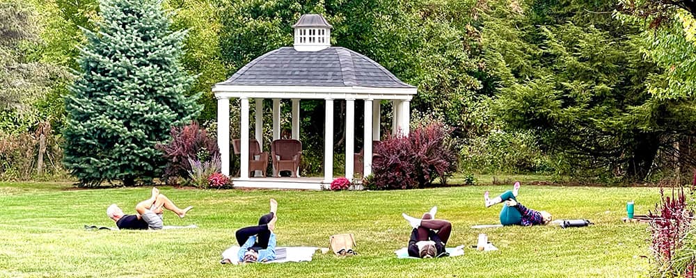 Five people practice yoga on a grassy field in front of a white gazebo surrounded by trees.