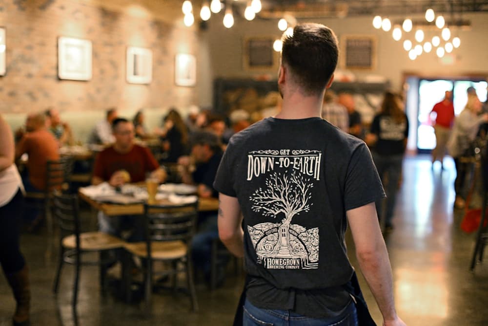 A server wearing a "Down-to-Earth" shirt walks through a bustling restaurant filled with patrons.