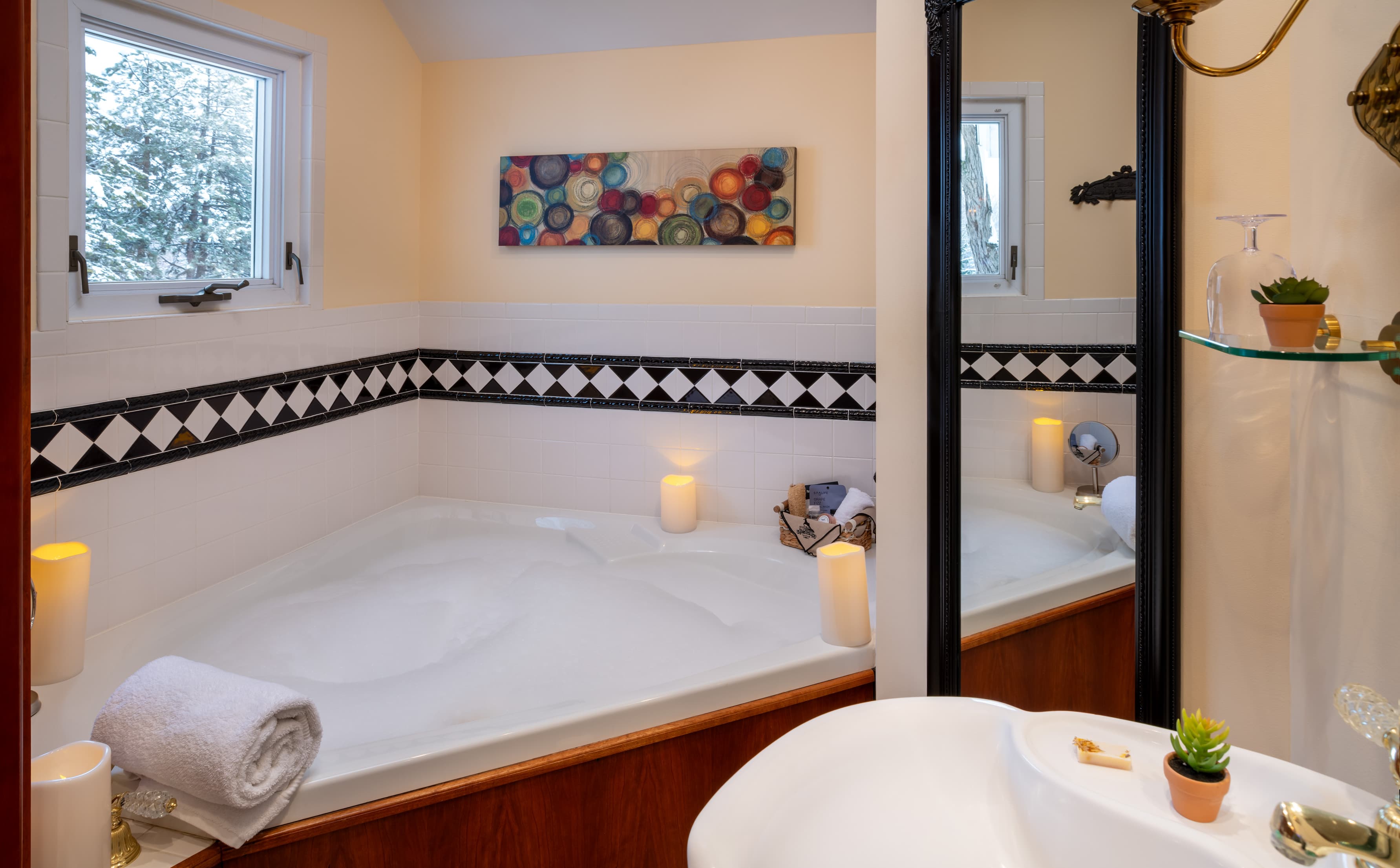 A relaxing corner jetted bathtub filled with bubbles, surrounded by glowing candles and a rolled white towel. The bathroom features cream-colored walls, a black and white diamond-patterned tile border, colorful abstract wall art, and a snowy view through a small window.