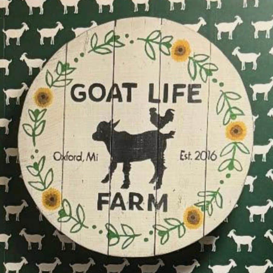 A decorative wooden sign reading "GOAT LIFE FARM" with a goat and chicken illustration, established in 2016, set against a patterned background of white goats.