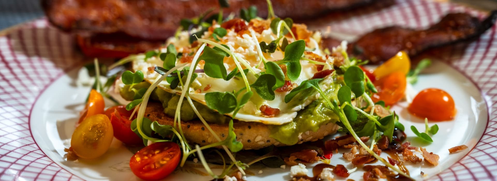 A plated dish featuring a bed of avocado, topped with a poached egg, crispy bacon, cherry tomatoes, and microgreens.