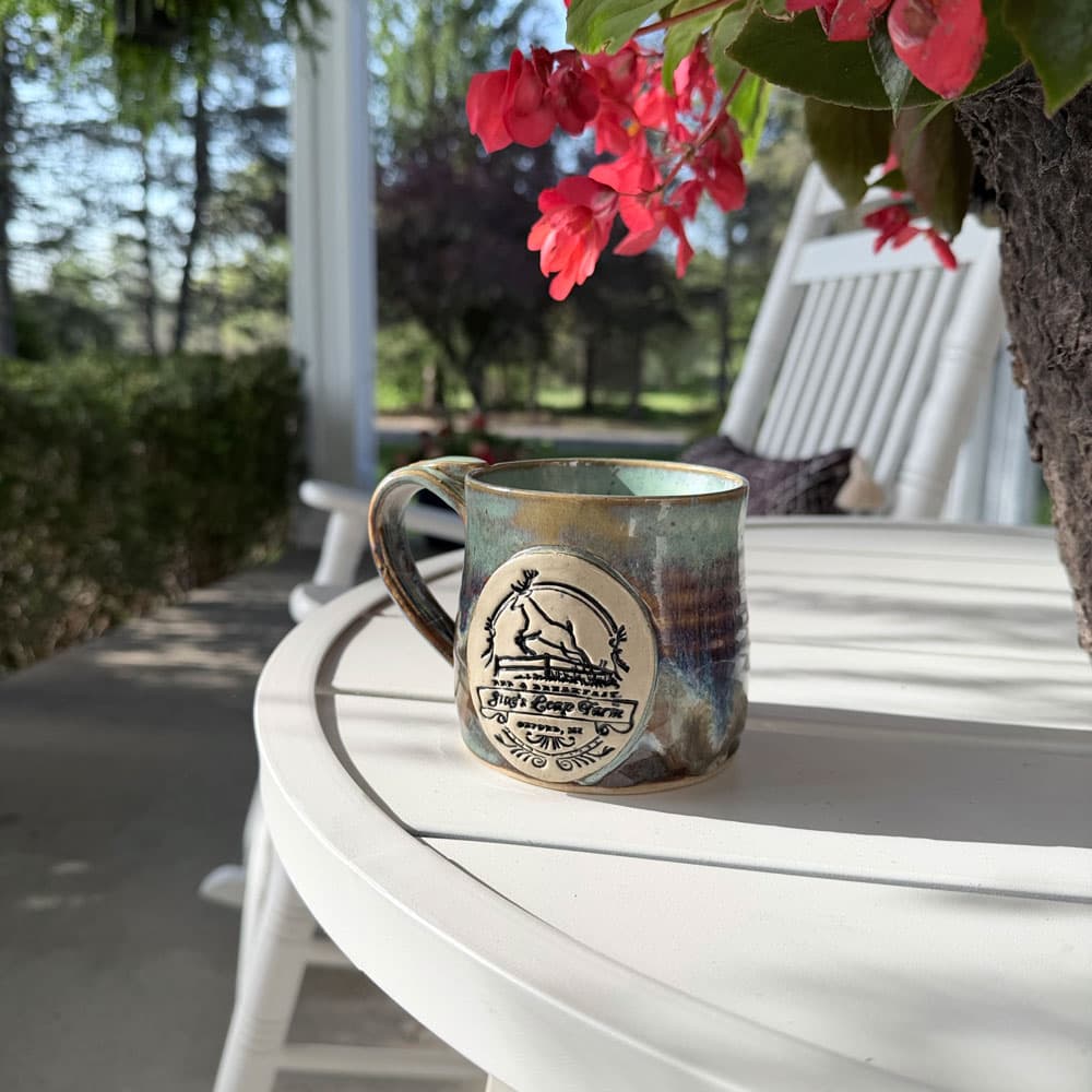A handcrafted mug with a decorative emblem sits on a white table surrounded by greenery and flowers.