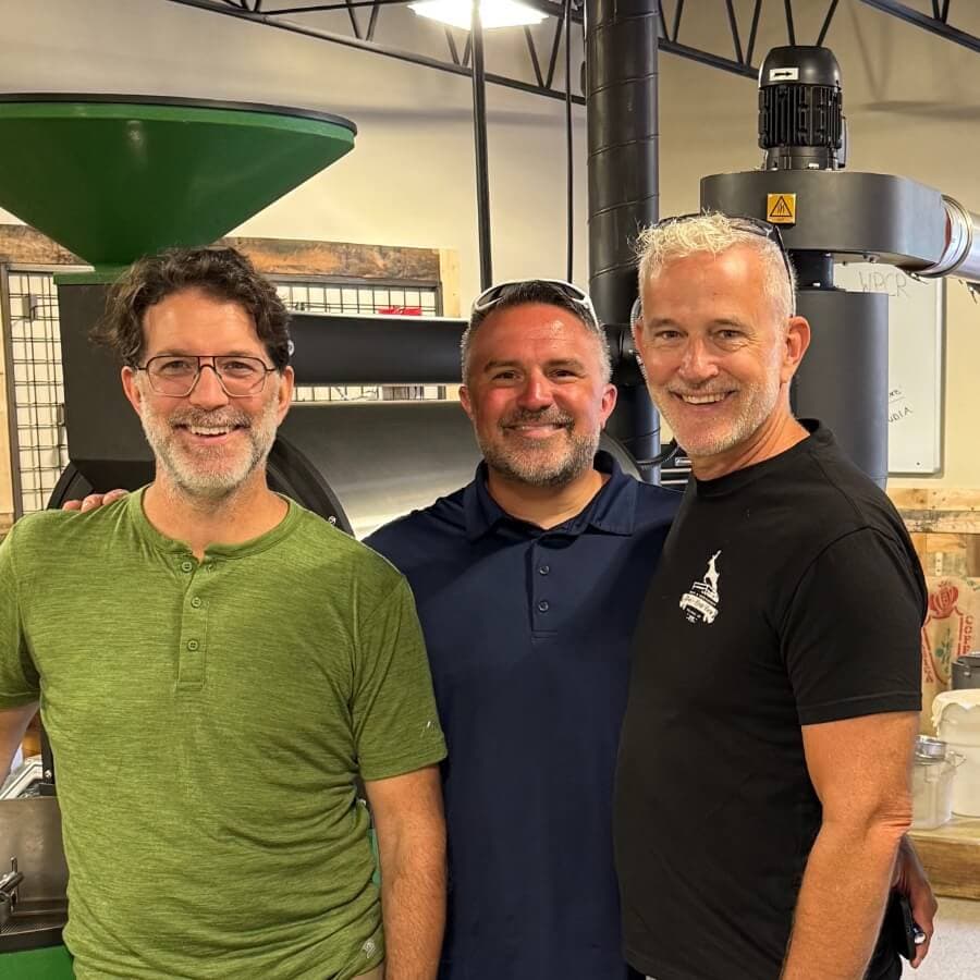 Three smiling men stand together in a coffee roasting environment.