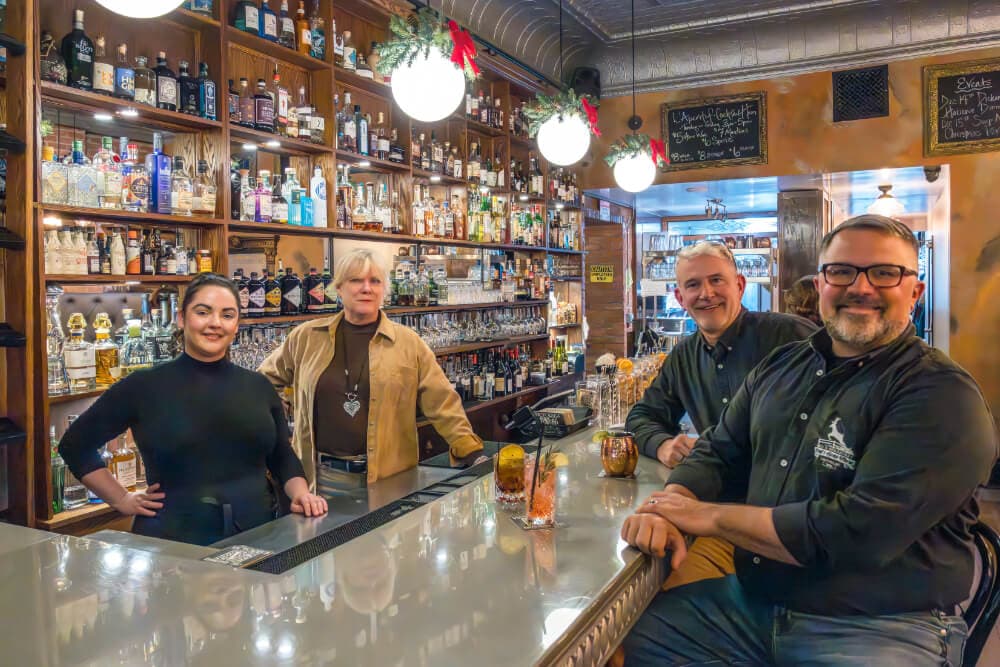 Four bartenders stand behind a polished bar filled with various bottles in a warmly lit establishment.