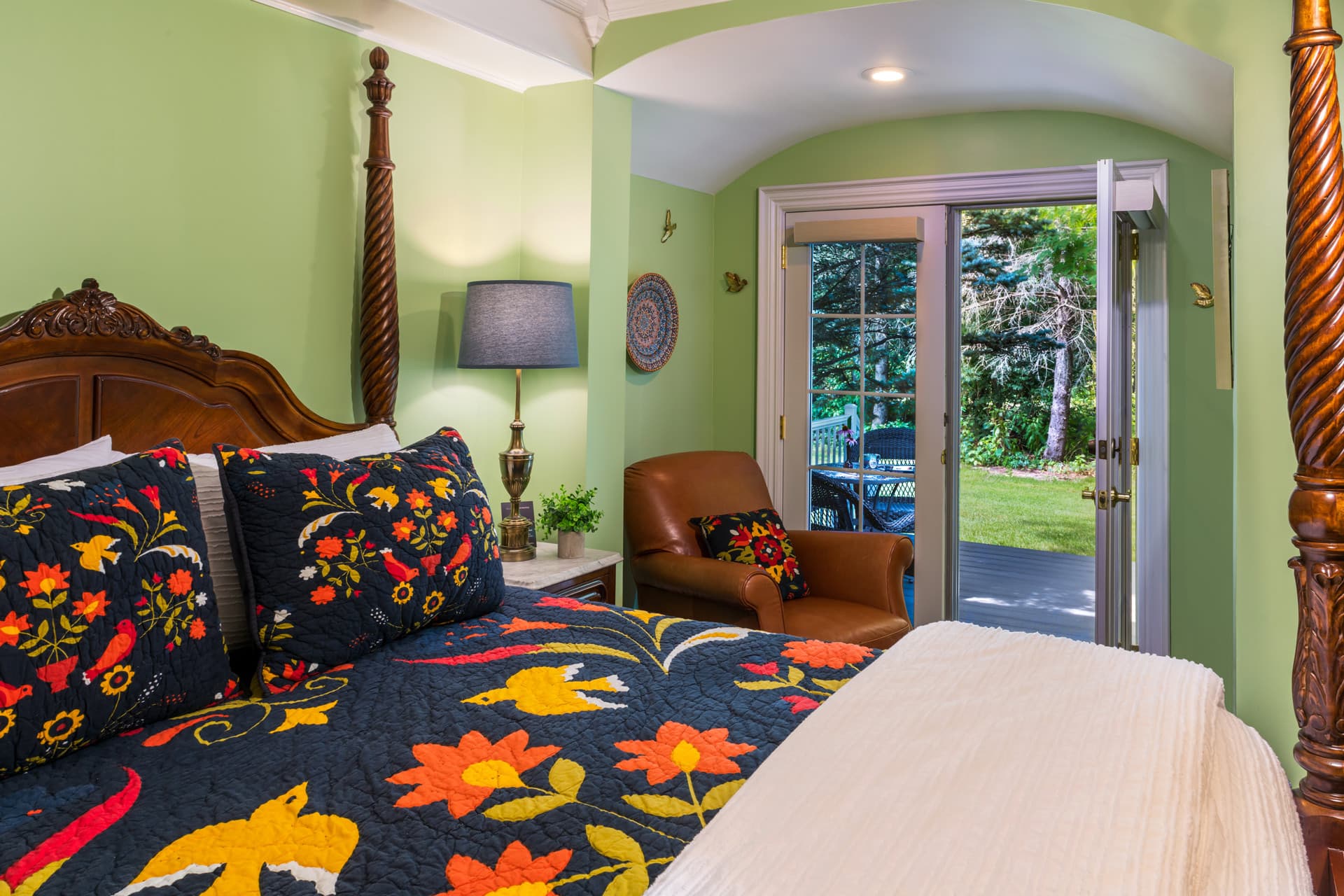 A vibrant bedroom with sage green walls and a wooden four-poster bed. The bed features a white quilt and dark blue pillows with colorful floral embroidery. In the background, a tan leather armchair sits near a set of glass doors leading to a snowy outdoor view.