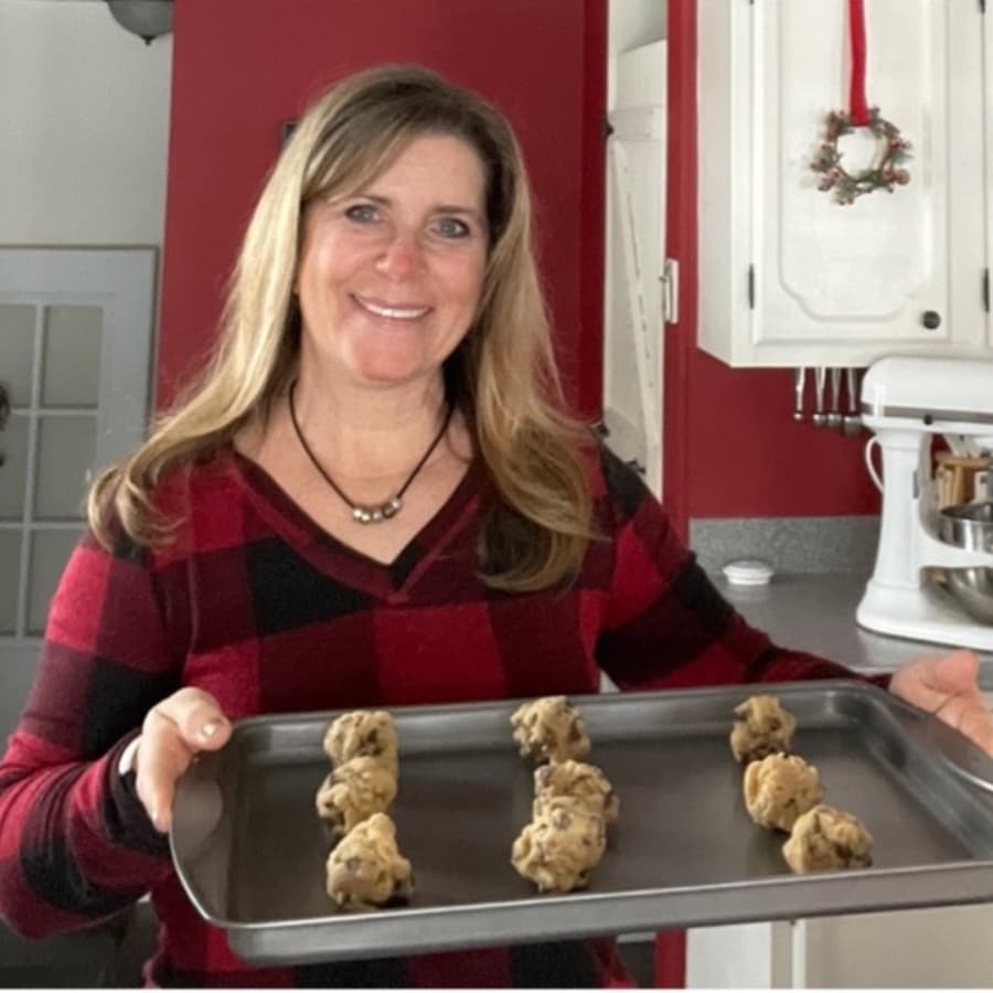 A smiling woman in a red plaid shirt holds a tray of cookie dough balls in a kitchen.