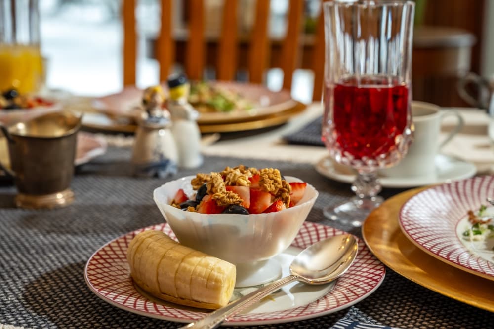 A breakfast table set with a fruit and granola bowl, a banana, and a glass of red beverage.
