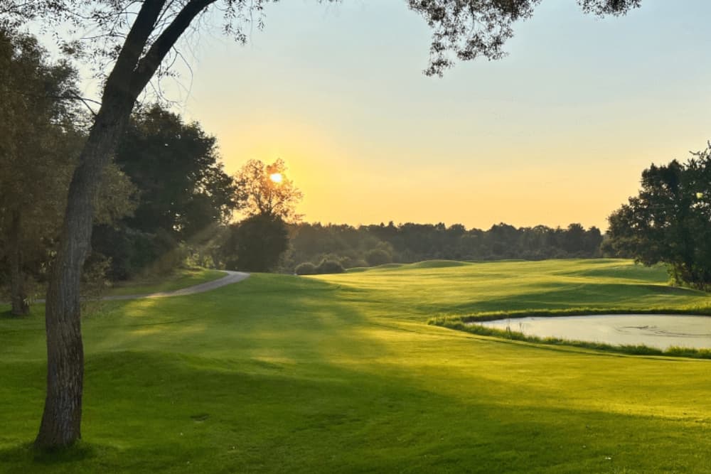 Sunset over a lush green golf course with a winding path and a pond.