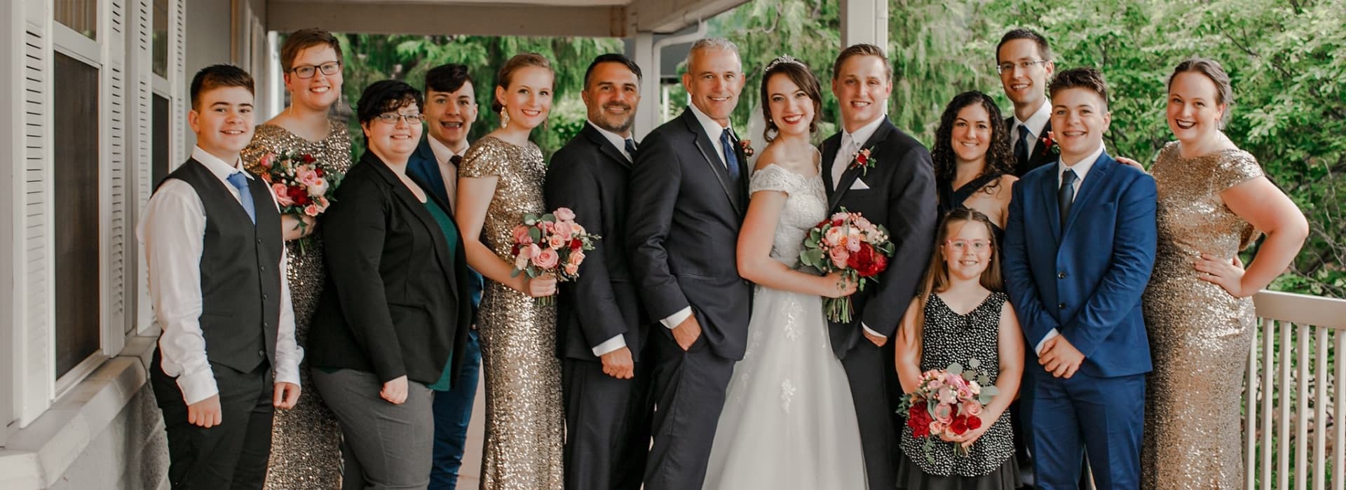 A large group of people, including a bride and groom, pose together on a porch, smiling and holding flowers.
