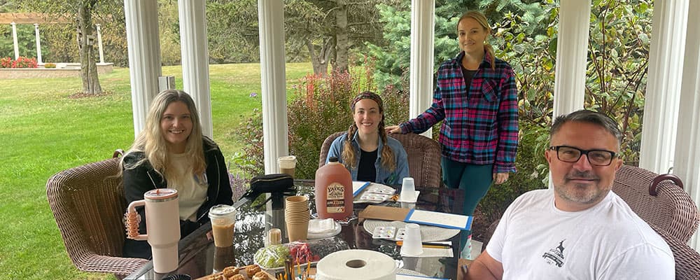 Four people are sitting around a table on a porch, enjoying food and drinks in a garden setting.