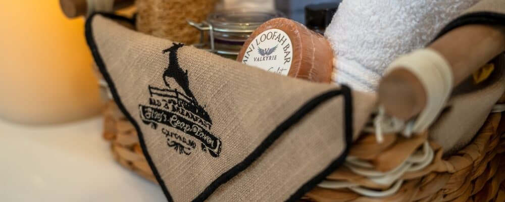 A woven basket filled with spa essentials, including a loofah, towel, and products, with a decorative burlap overlay featuring a logo.