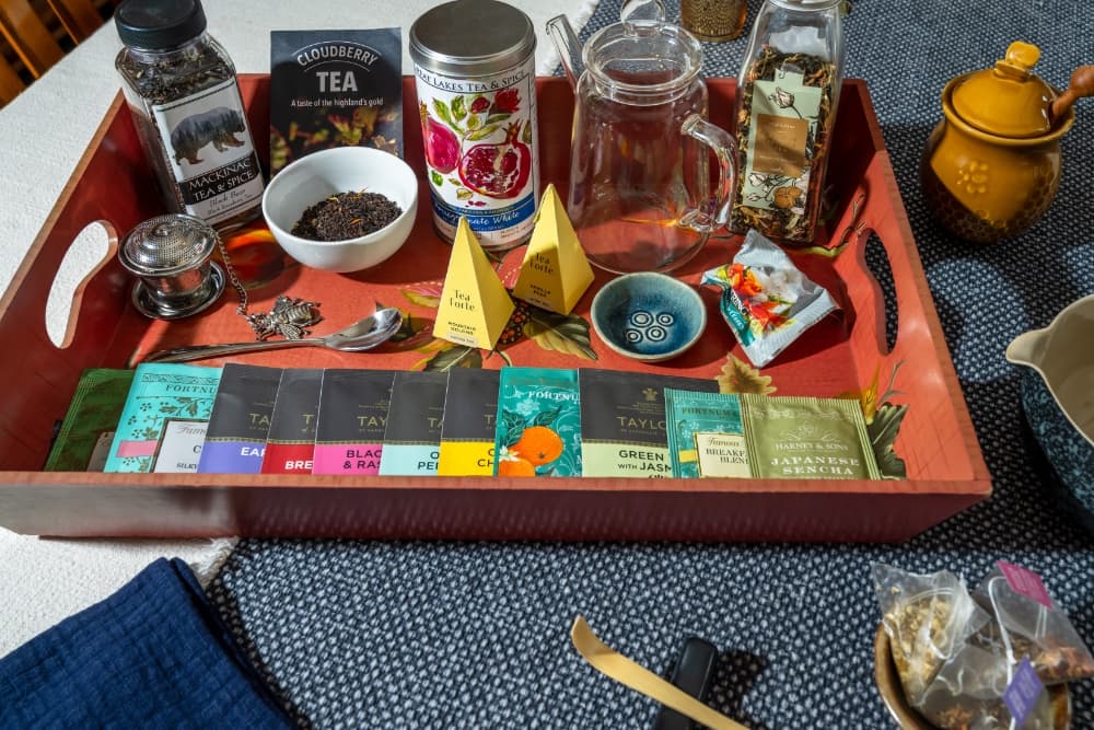 A wooden tray filled with assorted tea bags, a teapot, glass teapot, and various tea-related items.