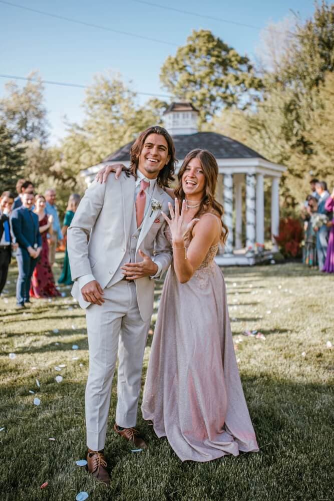 A man in a light suit and a woman in a pink dress smile and pose together outdoors, showing a ring on her hand.