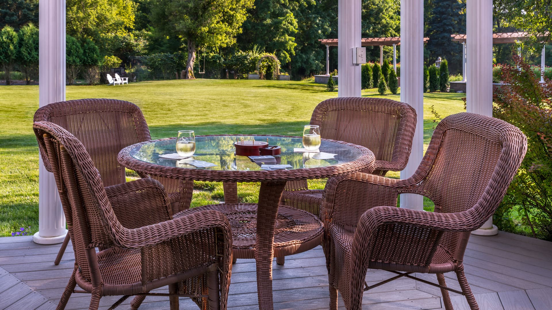 A cozy outdoor seating area with four wicker chairs around a glass-topped table, surrounded by a lush green lawn.