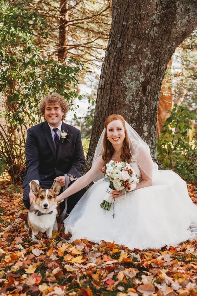 A bride and groom pose together with their corgi amidst colorful autumn leaves.