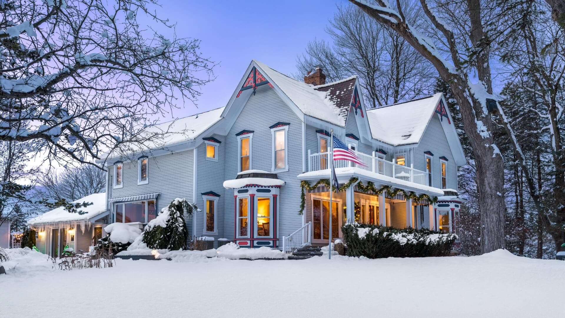 A quaint, snow-covered Victorian house adorned with festive decorations and an American flag.