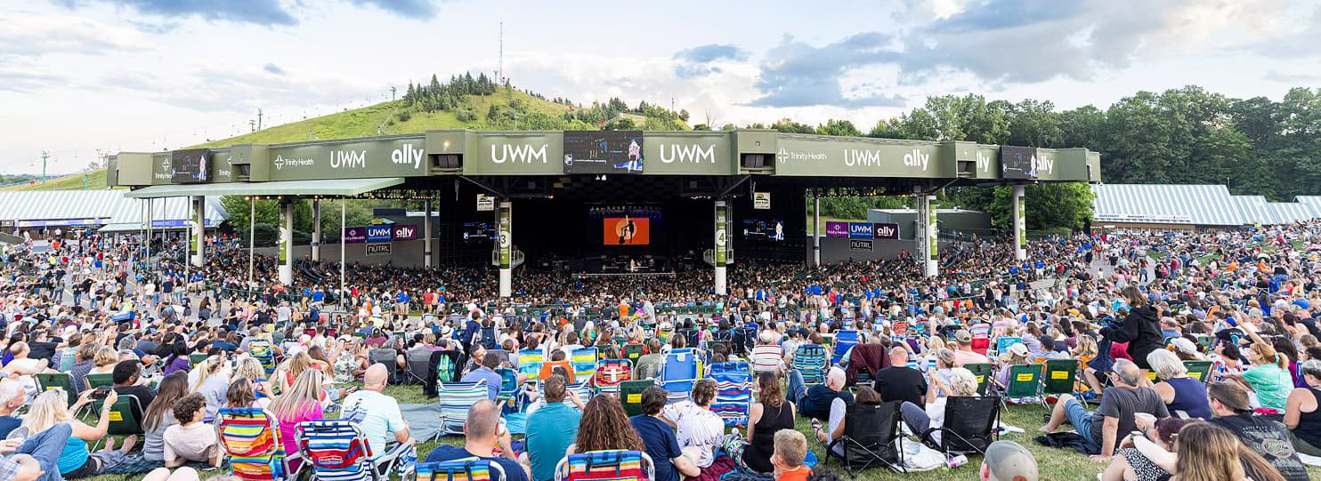 A large crowd enjoys a concert at an outdoor amphitheater surrounded by greenery.