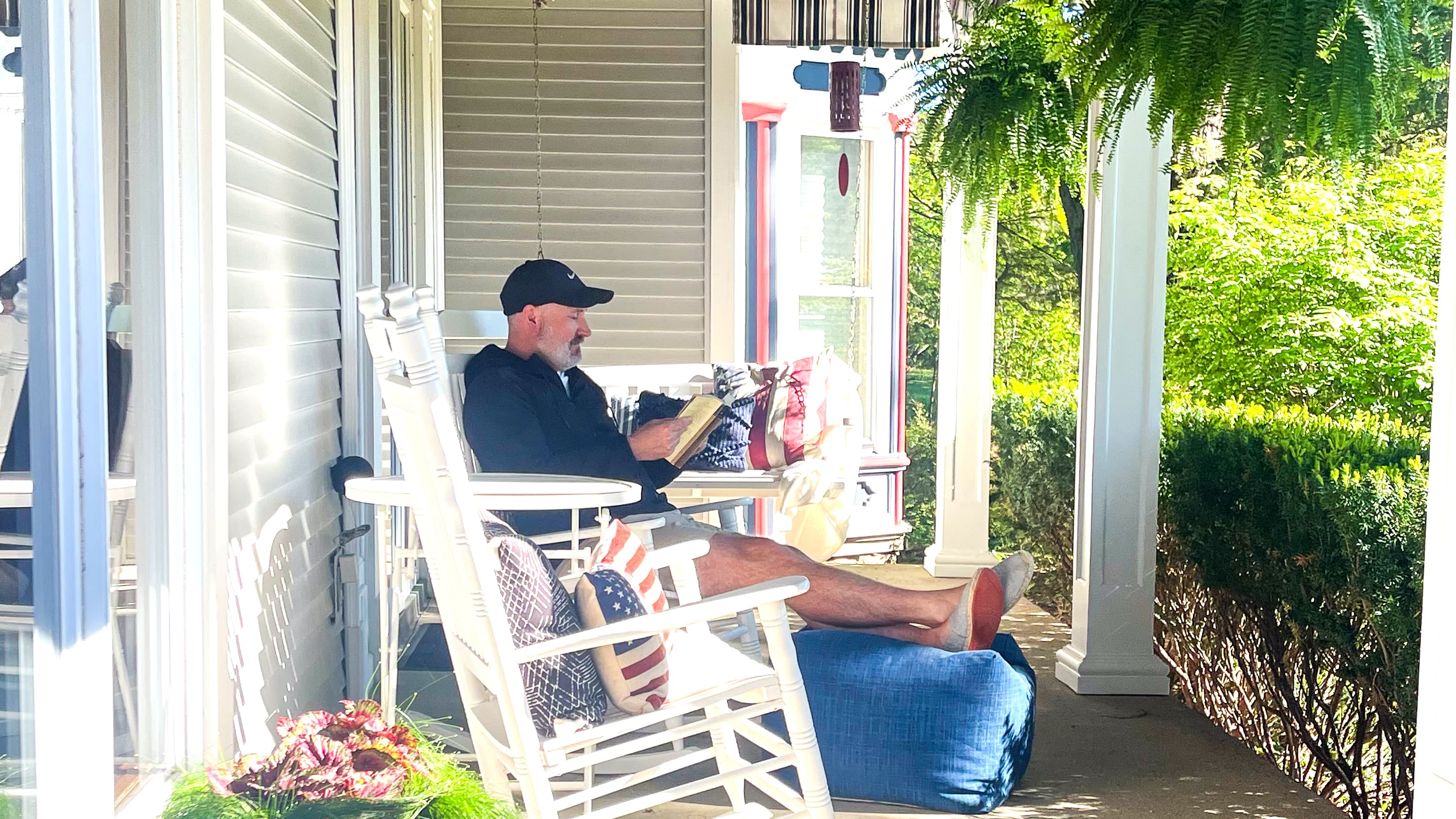 A man sits on a front porch in a rocking chair, reading a book surrounded by greenery.