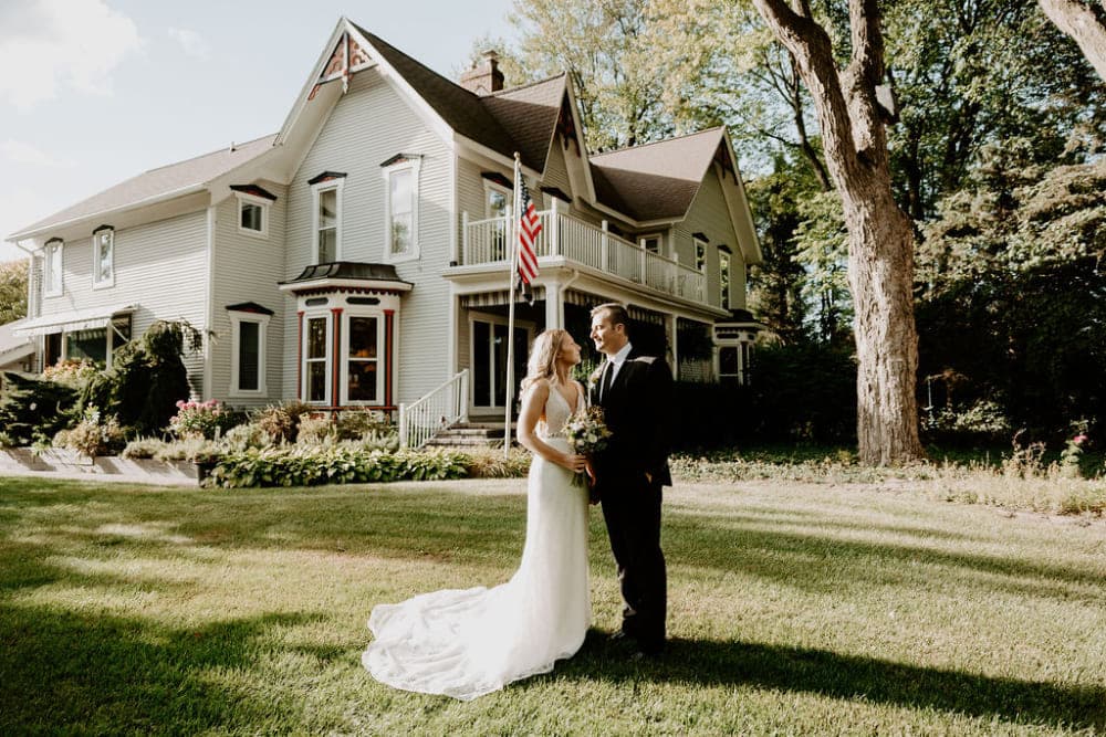 A bride and groom stand together in front of a large house, surrounded by greenery.