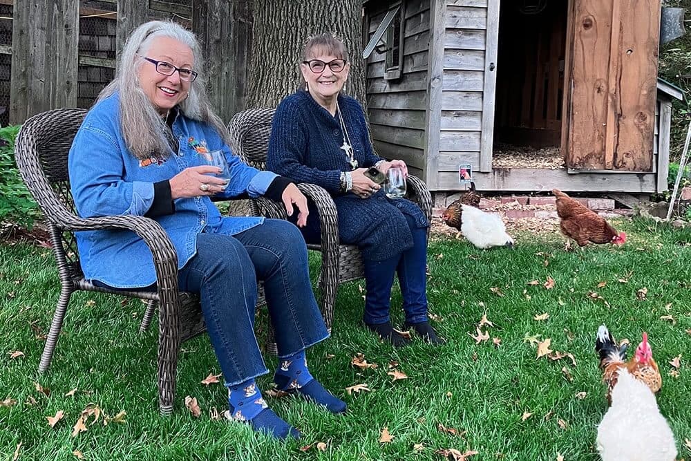 Two women sit on wicker chairs in a backyard with chickens roaming nearby.