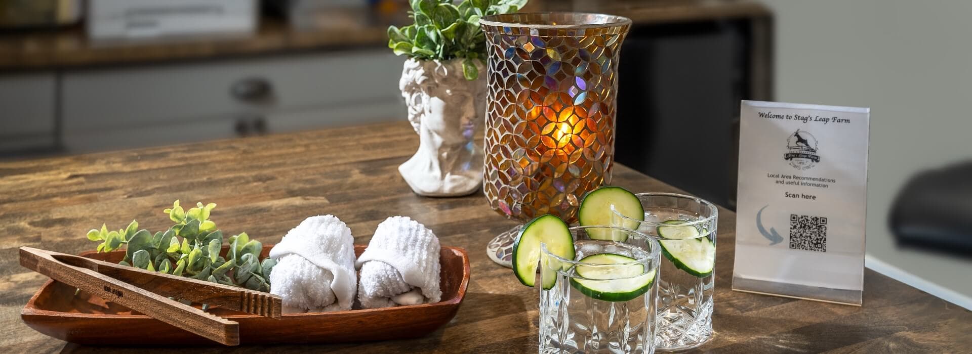 A wooden tray with garnished drinks, towels, and a decorative candle sits on a table alongside a welcome sign.