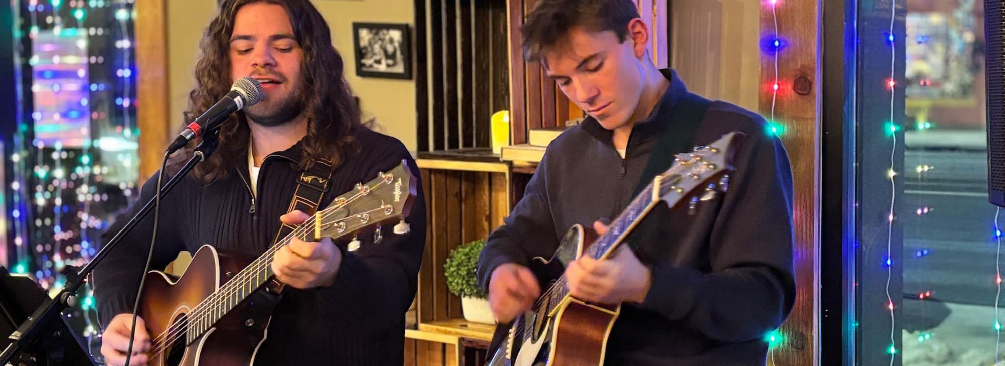 Two musicians perform with acoustic guitars in a festive setting illuminated by colorful lights.