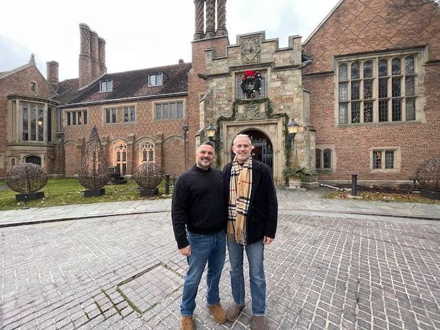 Two men stand in front of a large brick building adorned with holiday decorations.