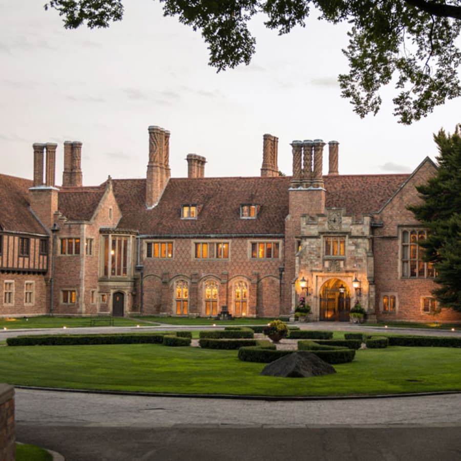 A grand brick mansion with multiple chimneys and large windows, set in a manicured lawn.