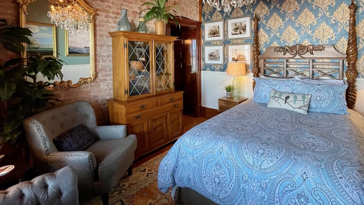 A cozy bedroom featuring a blue paisley bedspread, an antique dresser, and elegant decor against a patterned wallpaper backdrop.