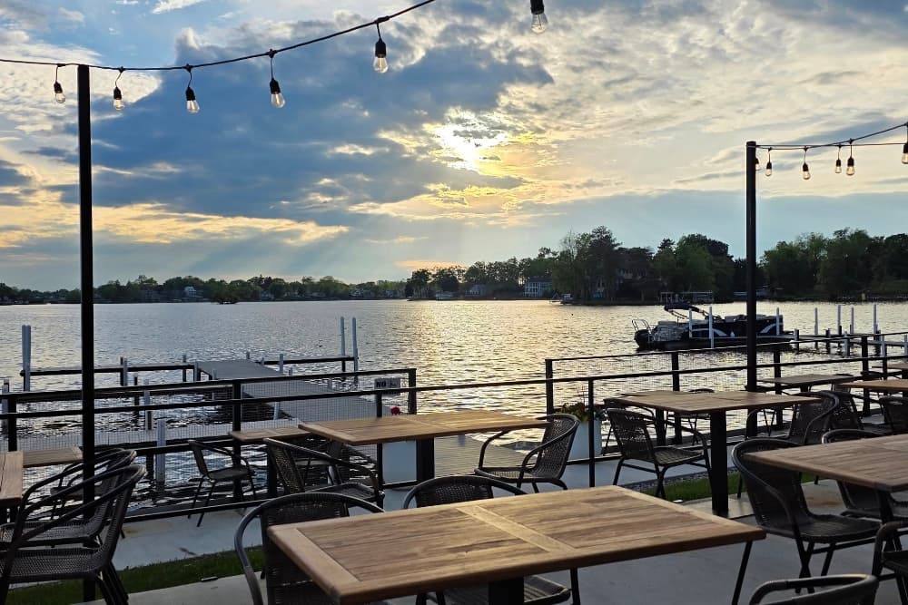 A lakeside dining area with empty tables and a sunset reflecting on the water.